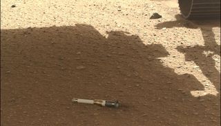 On a flat, slightly rocky ground, there is a sample tube laying there. A rover&#039;s wheel is seen toward the top right.