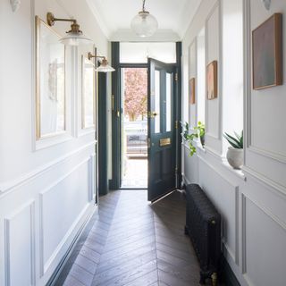 a hallway with open front door with white panelled walls and wooden herringbone flooring