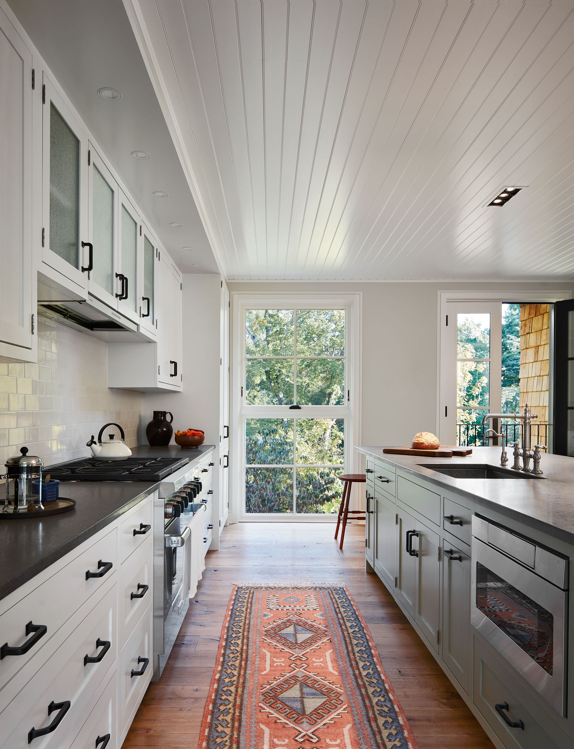 galley style kitchen with white units black worktops white paneled ceiling white tiled backsplash wooden floor and kilim rug blue island and full height windows and doors with treetop views