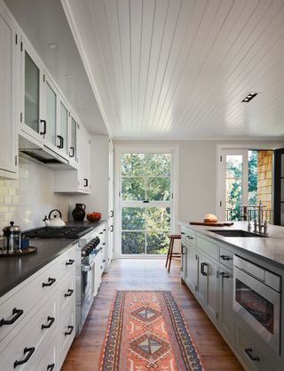 galley-style kitchen with white units black worktops white paneled ceiling white tiled backsplash wooden floor and kilim rug blue island unit and full height windows and doors with treetop view