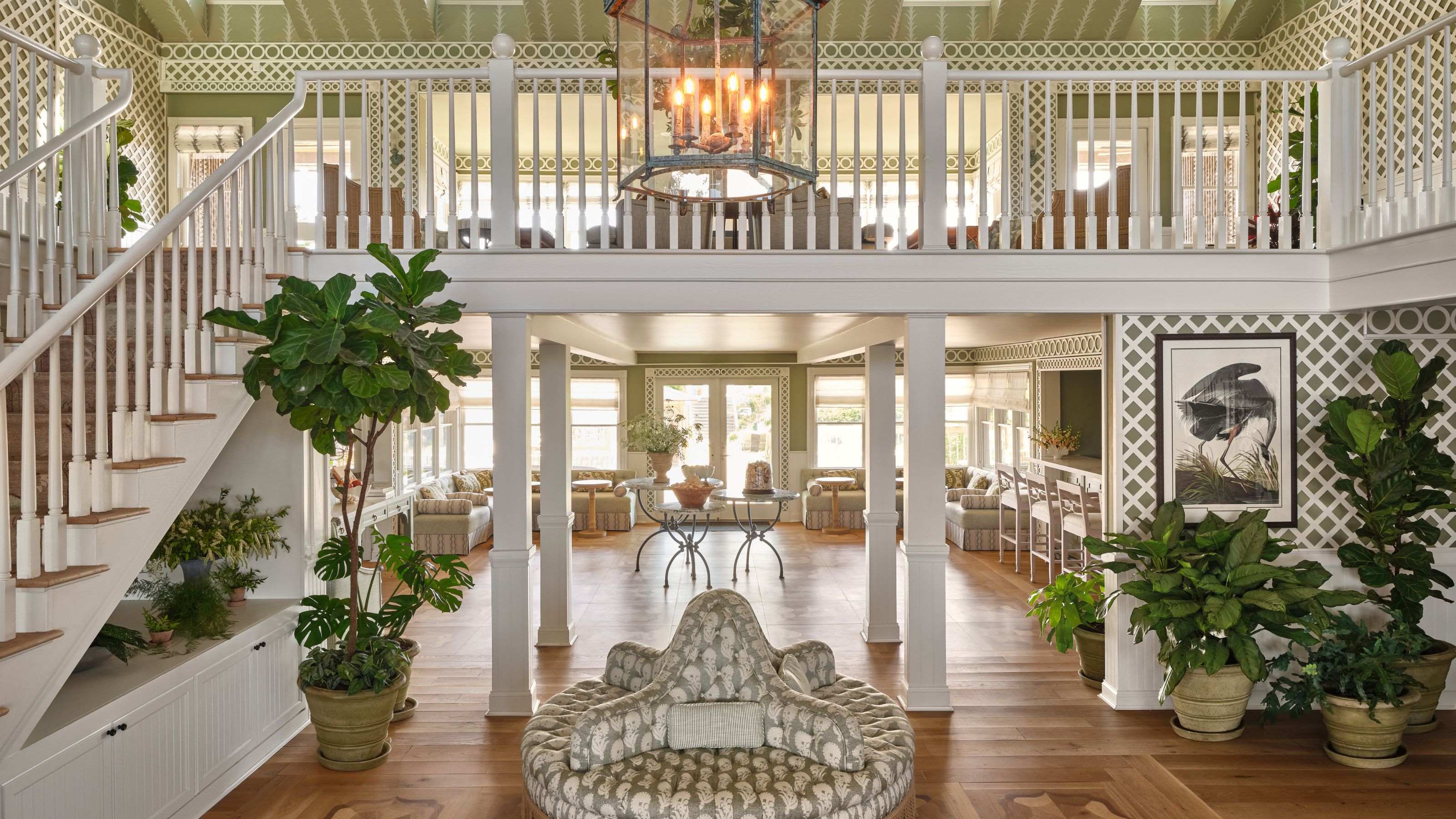 A spacious, two-story open-concept living area featuring white railings, a grand central staircase, green patterned accent walls, and vibrant potted plants throughout.