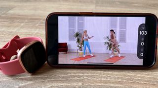 Smartphone showing Davina McCall workout on Fitbit Premium app, next to Fitbit Versa 4