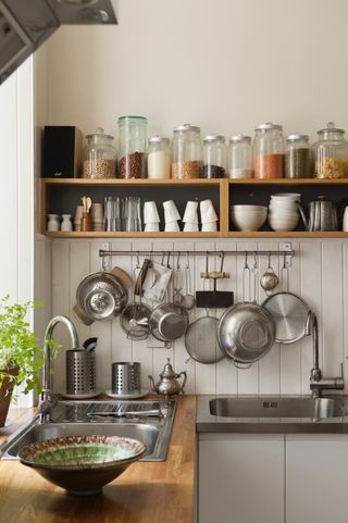 Dried ingredients on open shelving and utensils hanging from a pan rack in kitchen corner with sinks and solid oak work surfaces