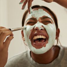 A woman in laughing while her friends (hands shown) are applying a green clay mask to her face