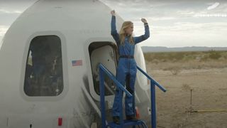 a woman in a blue flight suit raises her arms in triumph after exiting a white space capsule in the desert