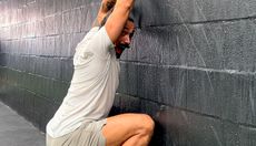Trainer Gustavo Vaz Tostes performing the wall-facing squat. His knees, toes and hands are in contact with a black wall, his knees are bent and his hips are below his knees. He has turned his face to the camera, his mouth is open and he has a friendly, triumphant expression.