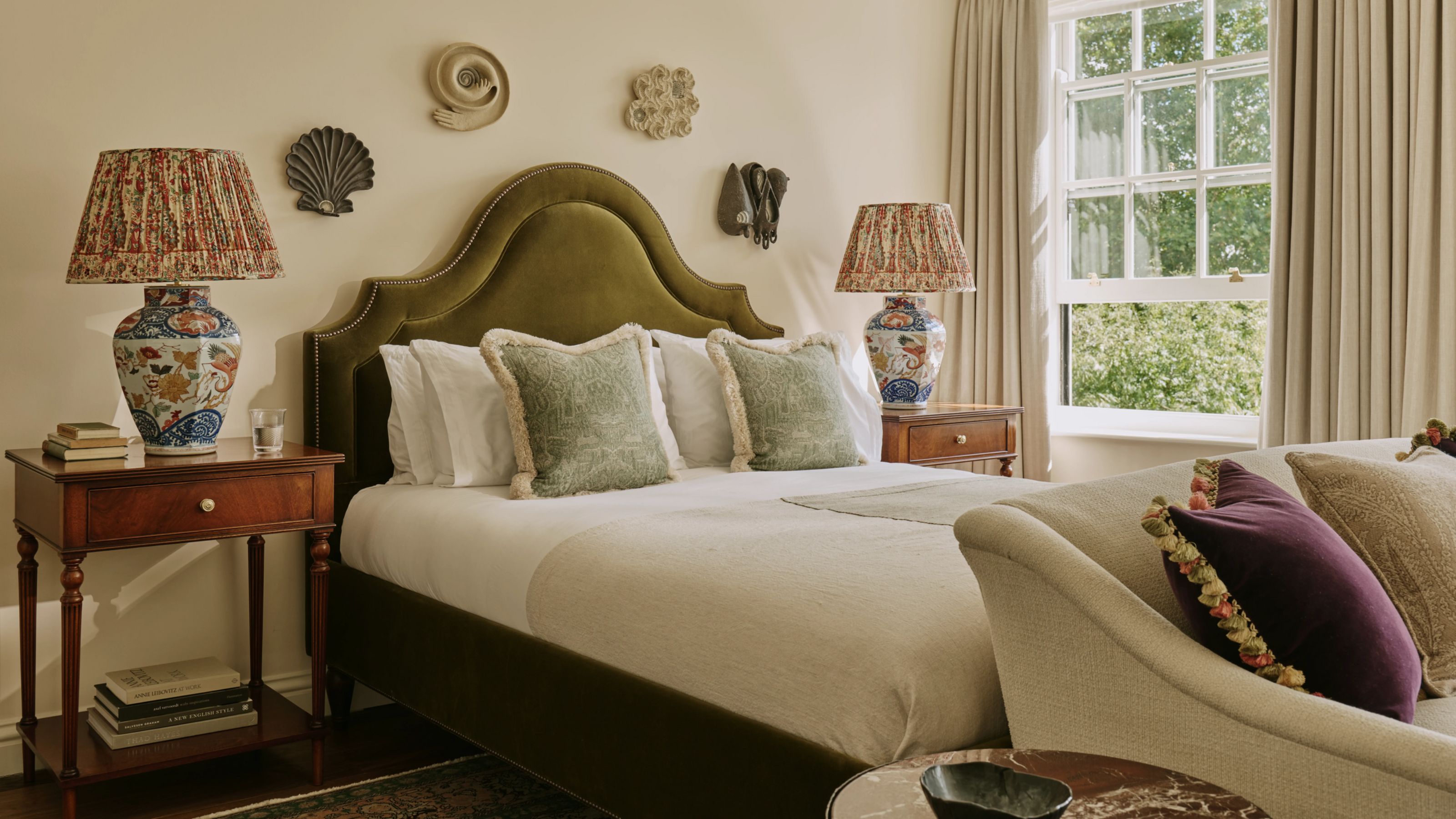 a soft warm neutral master bedroom with a green velvet headboard, two antique wooden side tables, vintage chinoiserie style lamps, and decorative plates on the wall behind the bed
