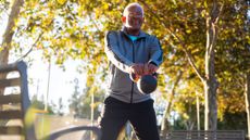 A man performs a kettlebell swing in a park. He is standing, legs straight, kettlebell grasped in his hands. His arms are also straight and the bell is swinging in front of him. Behind him we see trees and a bench.