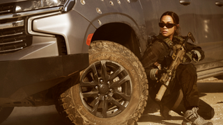 Zoe Saldaña as CIA operative Joe, wearing dark glasses and wielding a weapon as she ducks down beside a bullet-hole riddled van in Lioness season 2