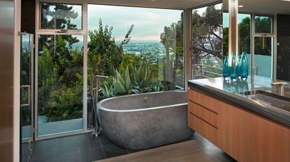 An earthy bathroom with a glass window and a tub with a concrete finish