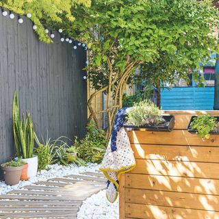 Corner of a garden with wooden stepping stone path, raised planters, a dark painted fence with festoon lighting attached