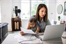 A woman sat in front of a laptop and paperwork with a baby on her lap