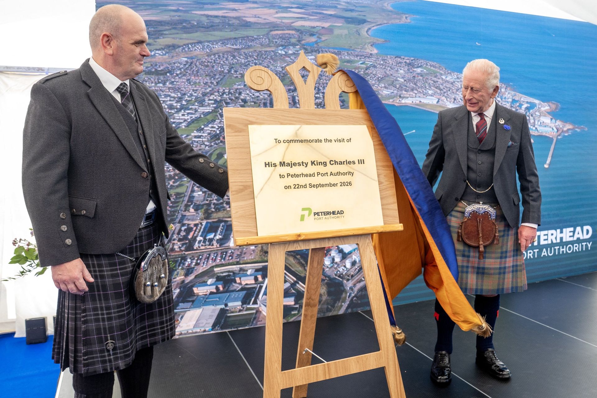 King Charles pulling a curtain off a plaque while wearing a kilt