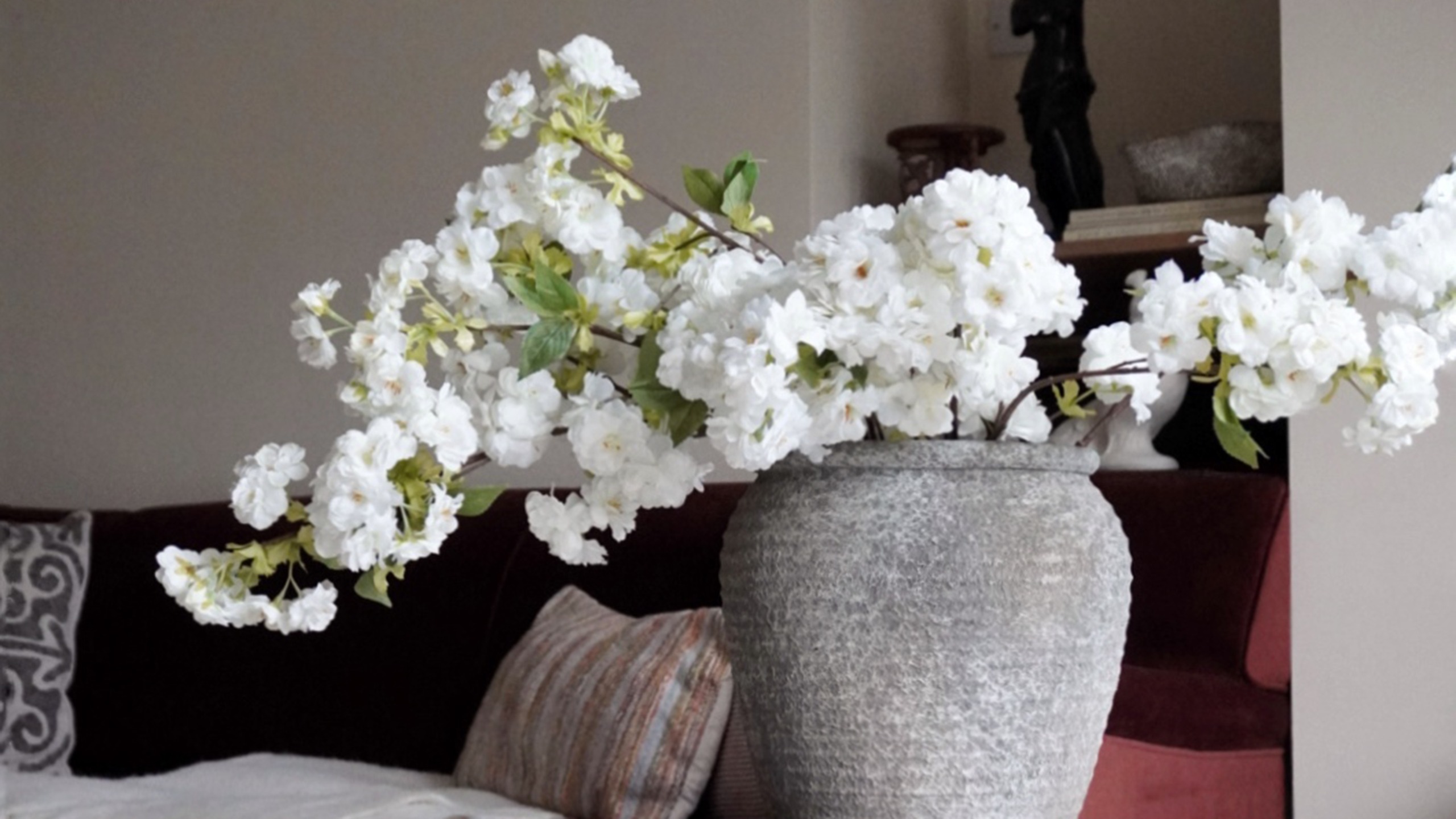 Large grey vase of white flowers beside burgundy sofa