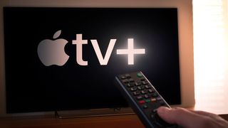 The Apple TV Plus logo on a TV in the background, while a hand holds a remote in the foreground