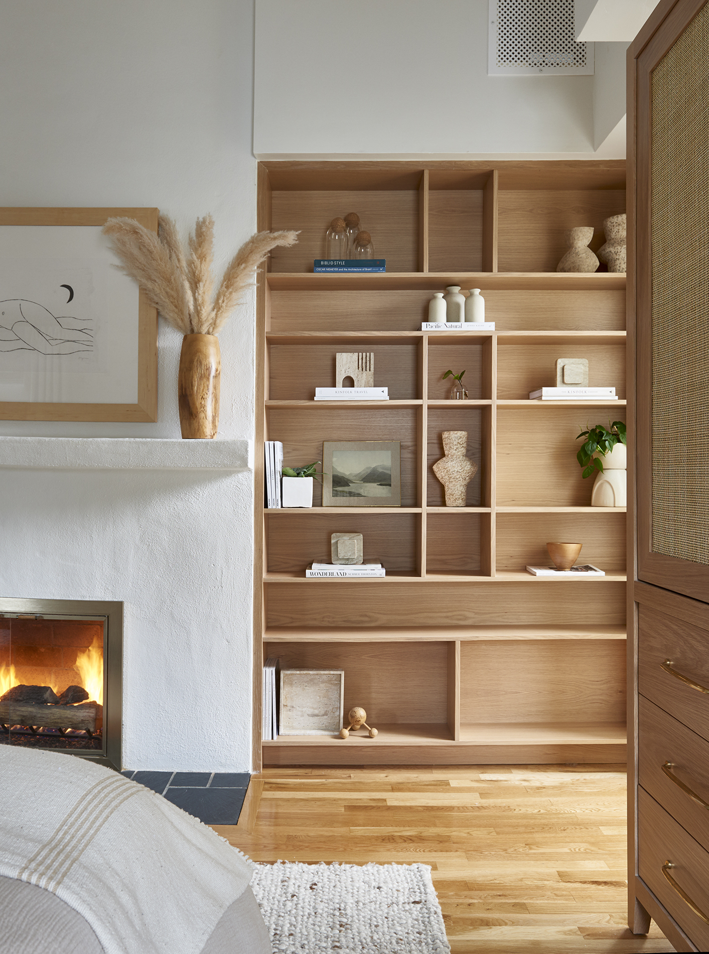 Image of a white, minimalist bedroom with custom wooden built-in bookshelves. There is a white plaster fireplace with a fire going.