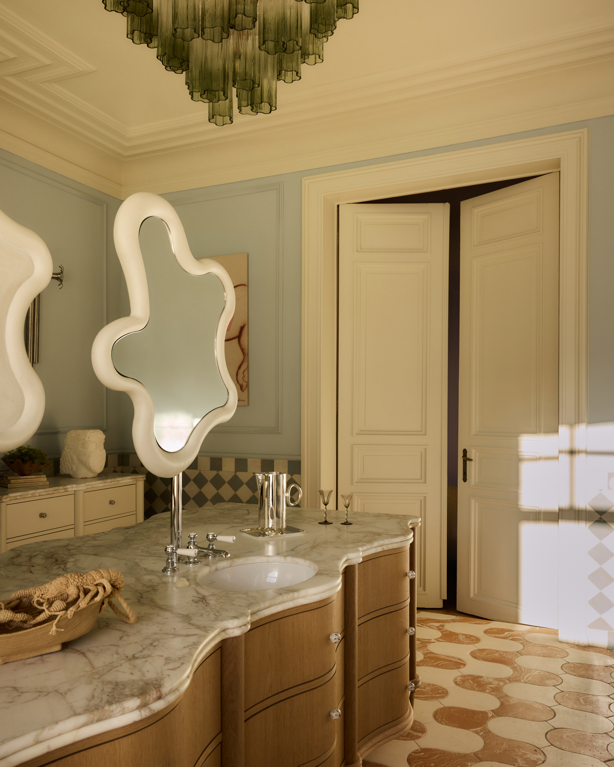 Image of a large bathroom painted light blue with white trim and a very unique tile floor. There is a wavy wooden island in the center with a marble top. There are suspended squiggly mirrors on the island.