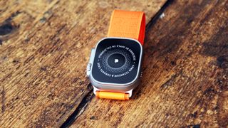 En Apple Watch Ultra med ett orange armband ligger utomhus på en träbänk.