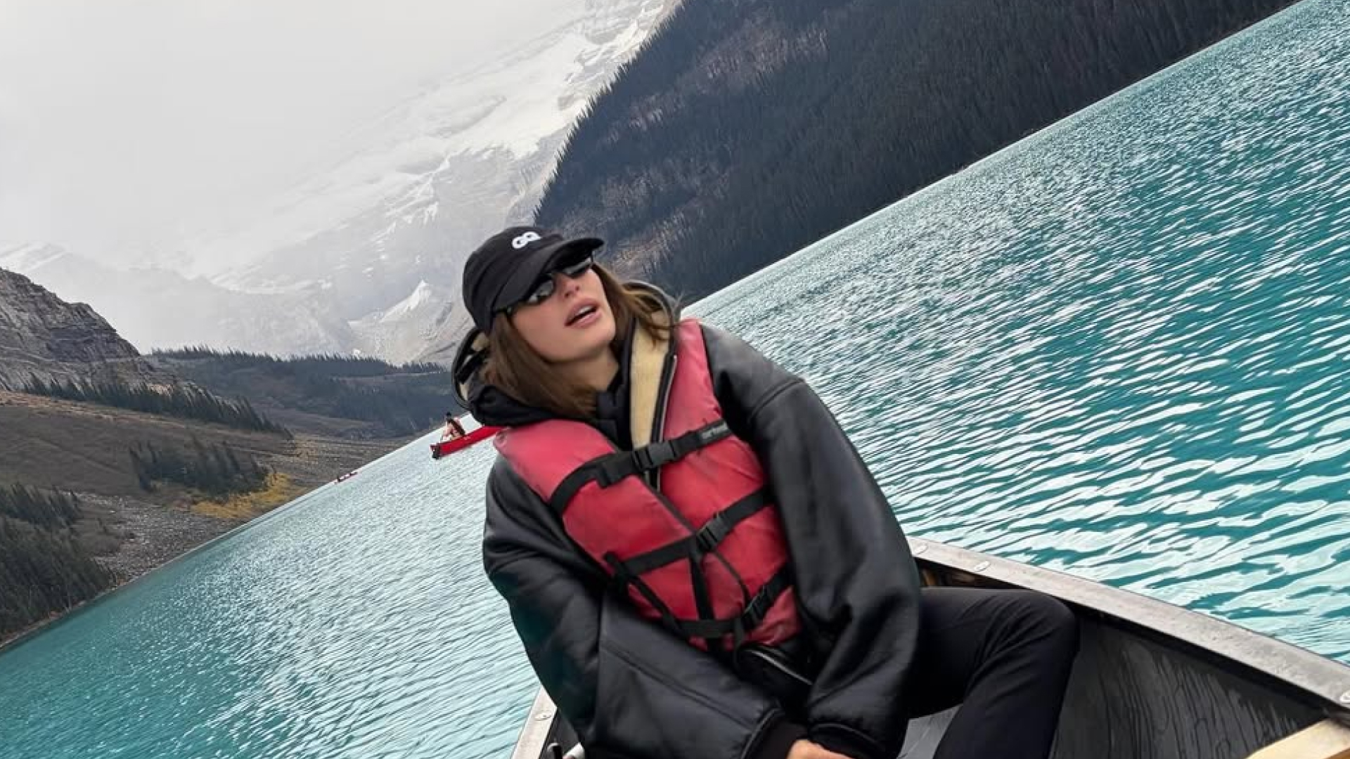 Hailey Bieber wearing a shearling jacket, baseball cap, and sitting in a lake