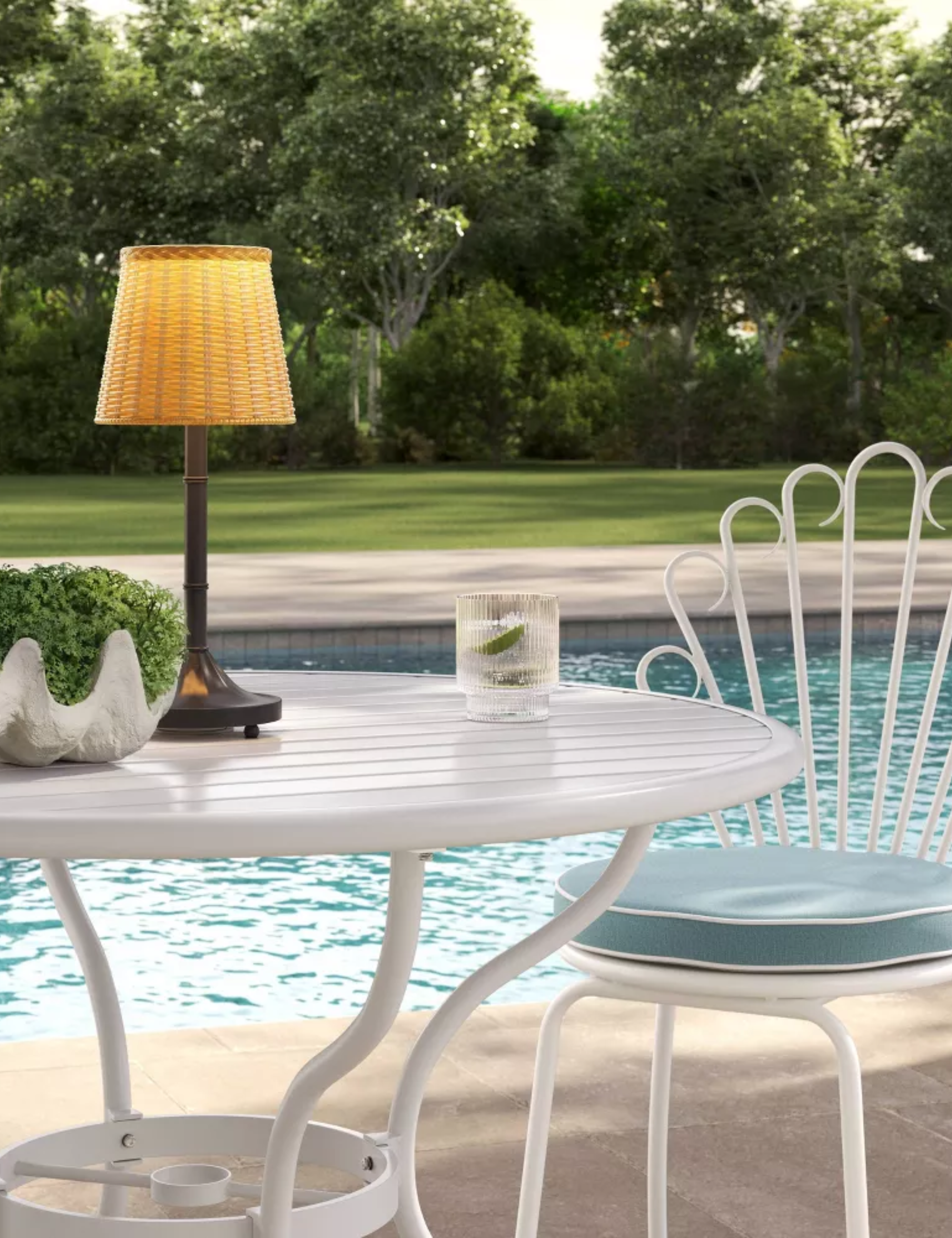 Portable wicker outdoor lamp aglow atop of a white patio set overlooking a pool and grassy lawn