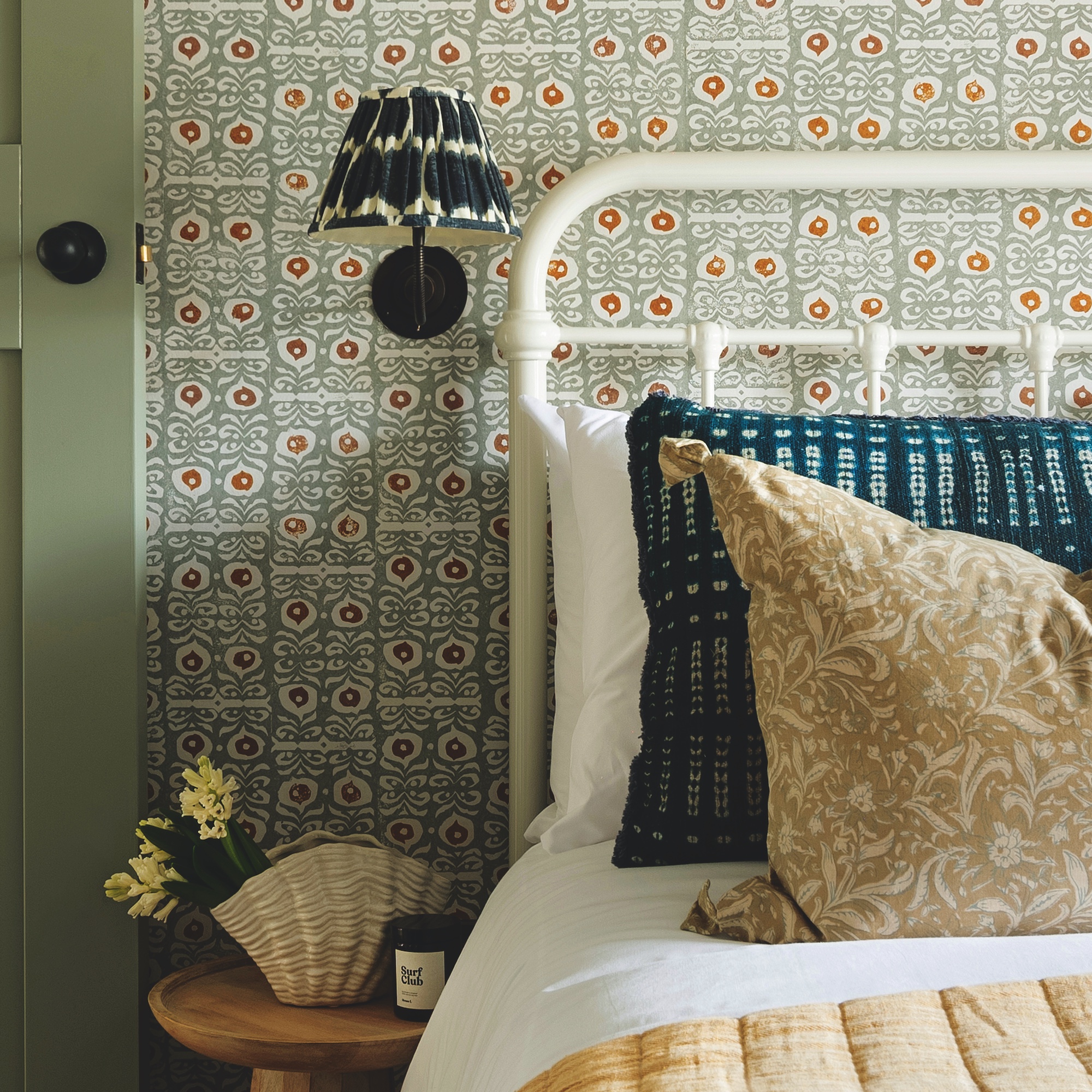 bedroom with small print wallpaper, white metal bedframe and wall light with ikat lampshade