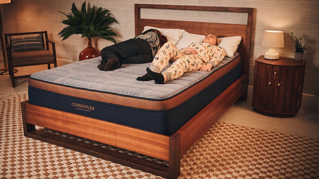 Two women lie on a Brooklyn Bedding CopperFlex mattress on a wooden bed frame. One woman in patterned pyjamas lies on her back, while the other in a black and white patterned shirt lies on her side facing away. The mattress features a textured blue-grey top, copper border, and dark blue base. The room is decorated with a checkered rug, a wooden nightstand with a lamp, and a potted plant in the background.