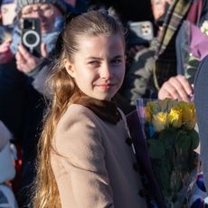 Princess Charlotte wears a light brown coat on Christmas Day in 2025 and is handed yellow and red flowers by royal fans in Sandringham