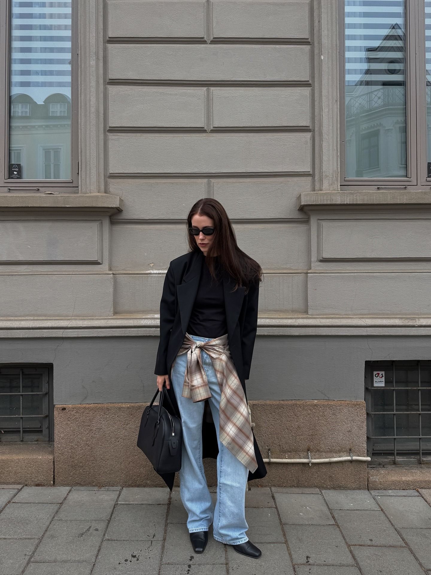 Influencer Annabel Rosendahl is in pale blue jeans, a black top, a black coat, and a plaid button-down tied around the waist.