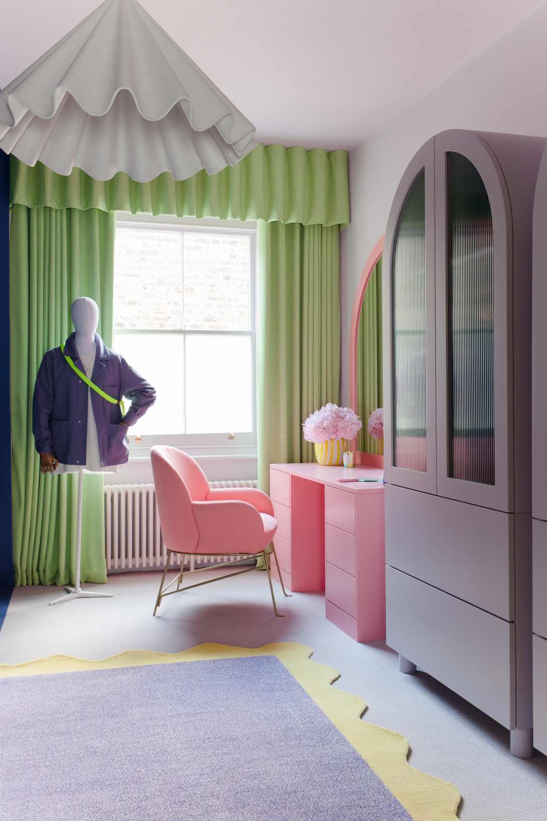 colorful room with purple hutch, lime green curtains with a valance, purple and yellow scalloped floor rug, pink vanity with matching pink chair and a vase of flowers, and a mannequin with a jacket and bag on