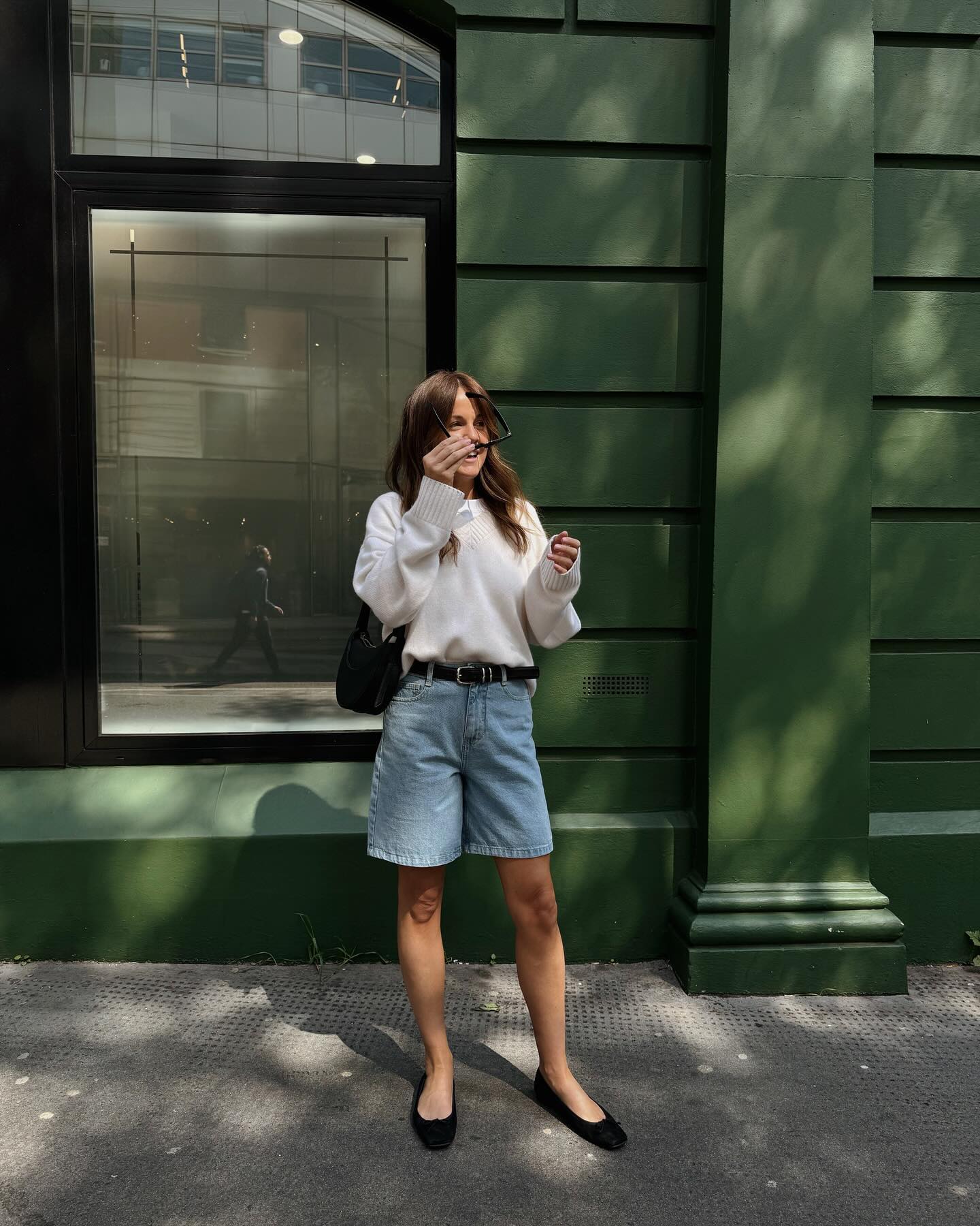 Woman on street wears white sweater, blue long shorts and ballet flats