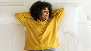 A woman in a yellow long-sleeved top lies on top of a white mattress and pillows