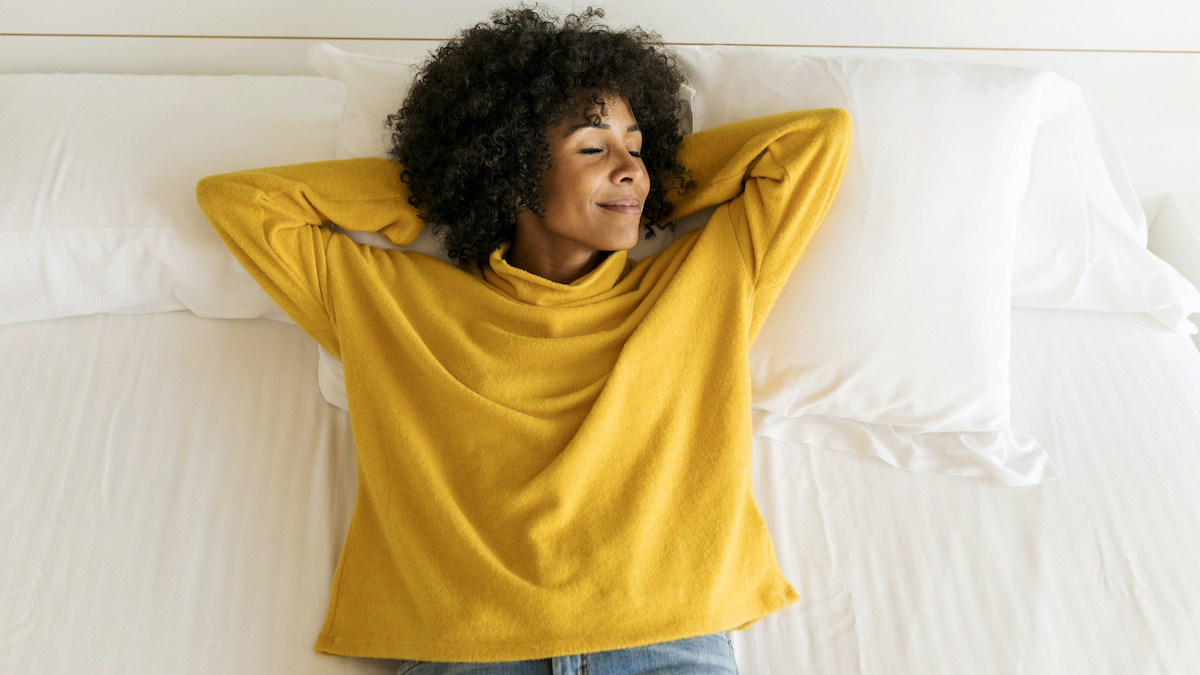 A woman in a yellow long-sleeved top lies on top of a white mattress and pillows