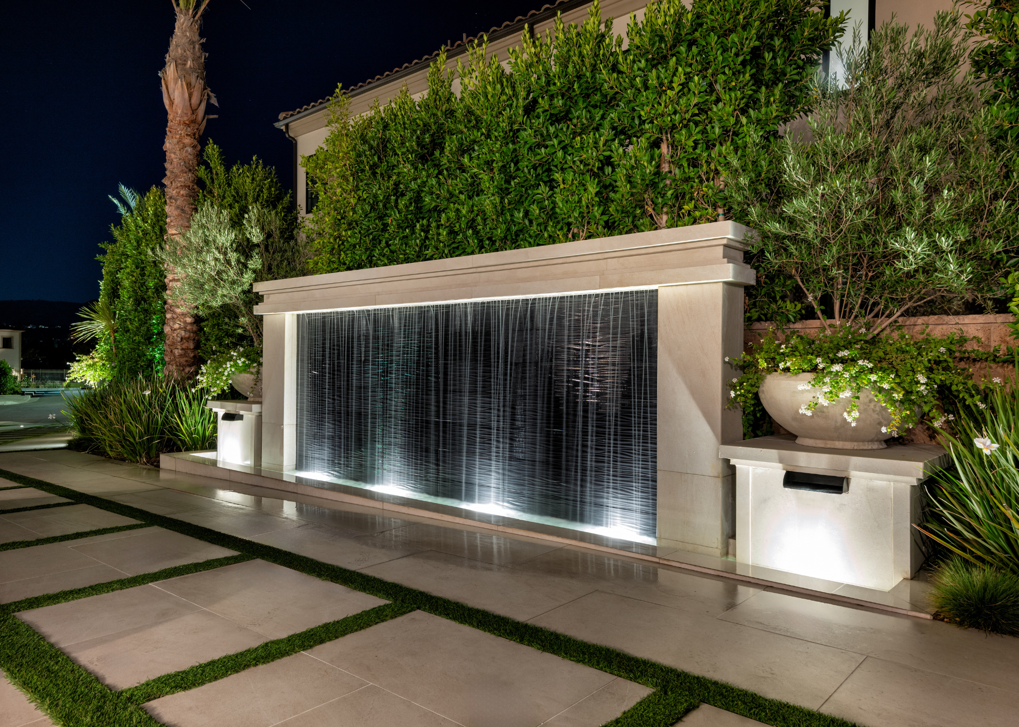 A modern California-style garden with a large, lit water feature surrounded by greenery in pots, beds, and up the walls of the sides of the house