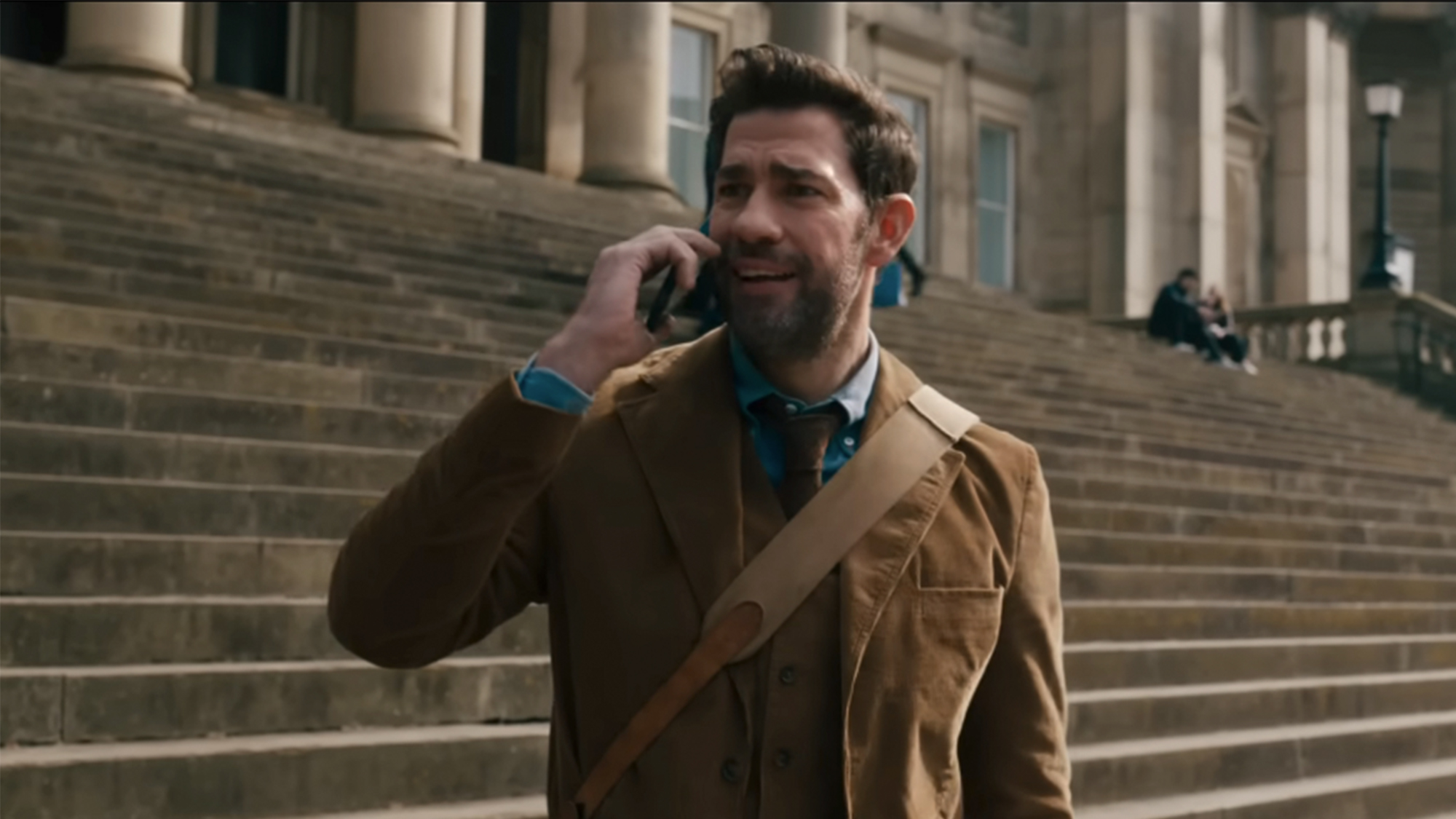 John Krasinski talking on a cell phone wearing a brown jacket in front of some stairs in Fountain of Youth