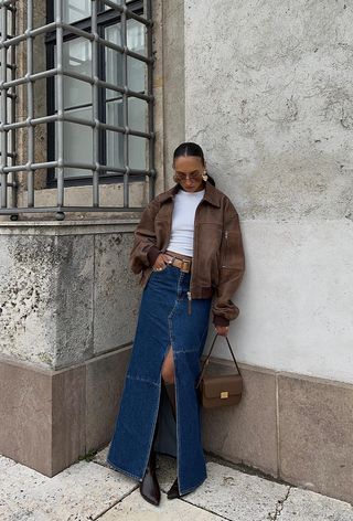 A denim skirt outfit is shown in a photo of a woman leaning against a wall outside wearing a brown leather jacket over a white t-shirt with a brown belt, denim maxi skirt, brown knee-high boots, a shoulder bag