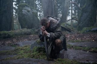 Charlie Hunnam as King Arthur kneels while holding his sword in a forest in King Arthur Legend of the Sword