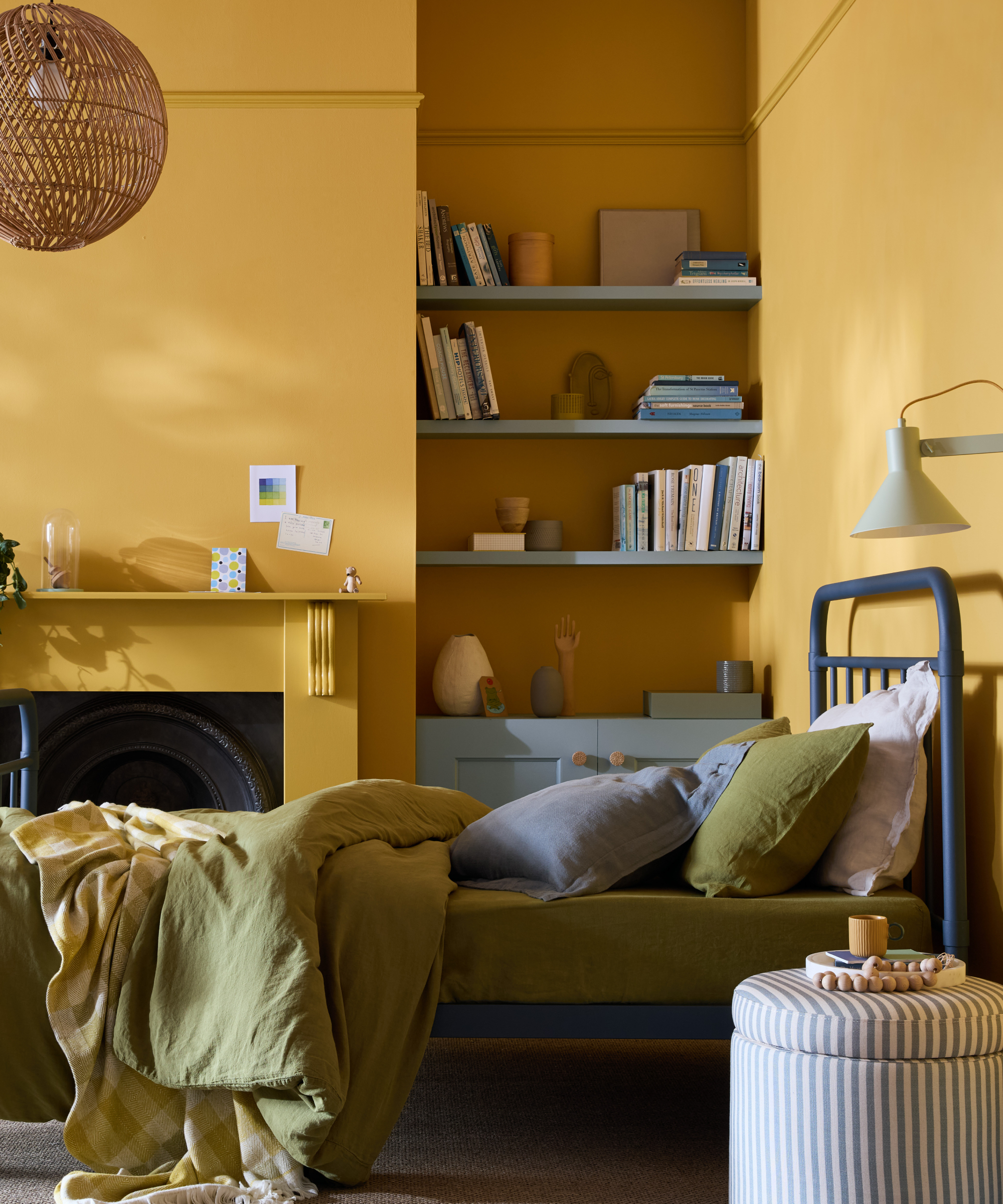 bedroom with yellow walls and fireplace, pale blue shelving and cupboard in alcove, blue bedframe with green and blue bedding