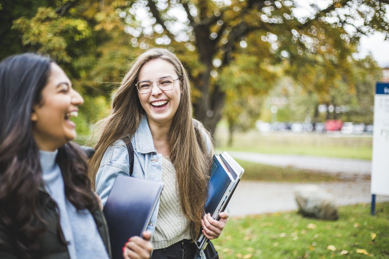 Two happy students walking outdoors together while carrying books and folders.