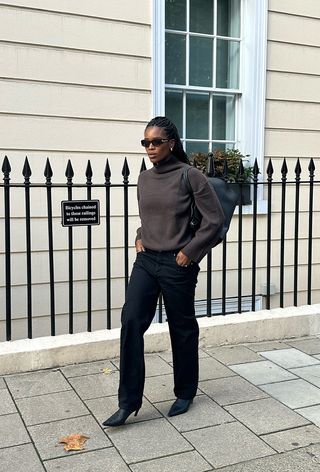 A woman standing outside wearing a brown turtleneck sweater with black straight-leg jeans, black kitten-heel ankle boots, a black bag, and brown sunglasses.