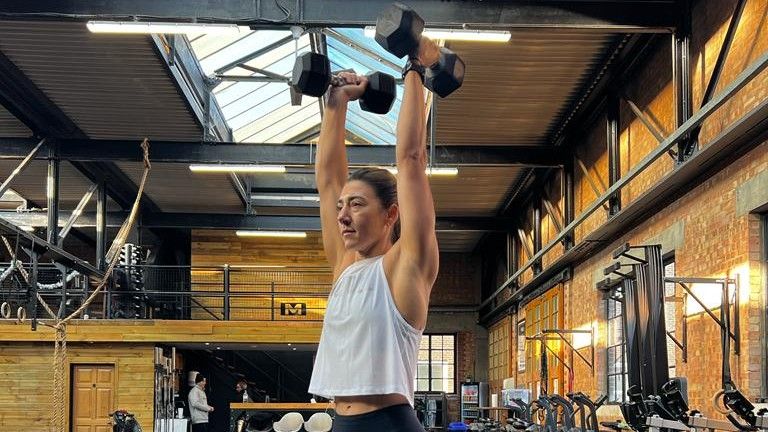 Jess holding both dumbbells extended above her head for shoulder press