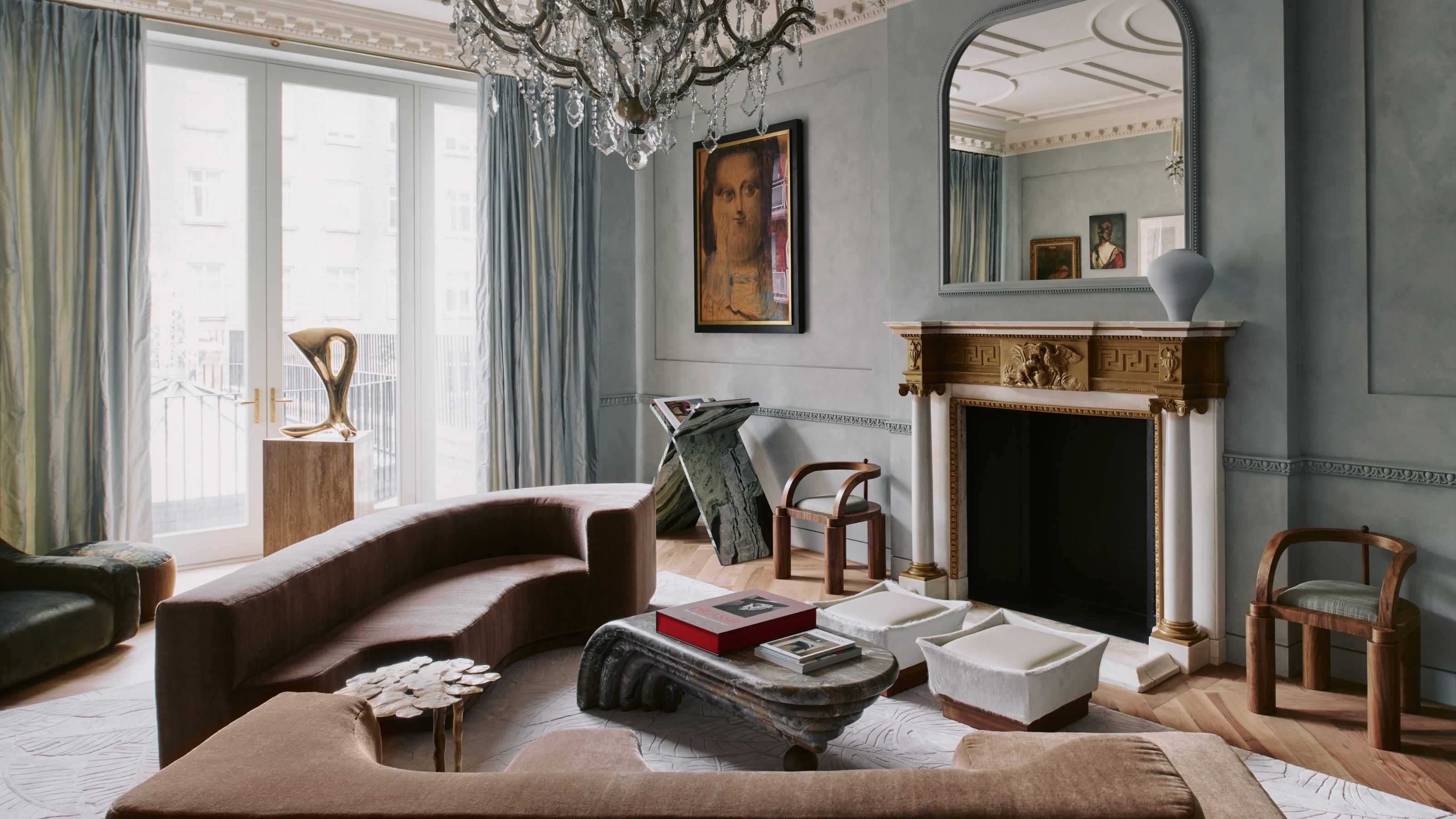 Image of a grand, light blue living room with a gold and white fireplace that has a mirror over it. There are two modern, brown sofas, two white pouffes, and a modern coffee table.