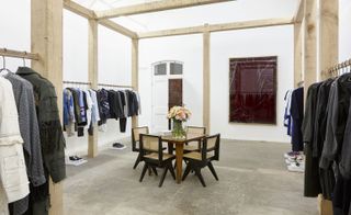 A clothing store with clothing rails and wooden pillars around the edges of the room and a square table with chairs in the middle of the room.