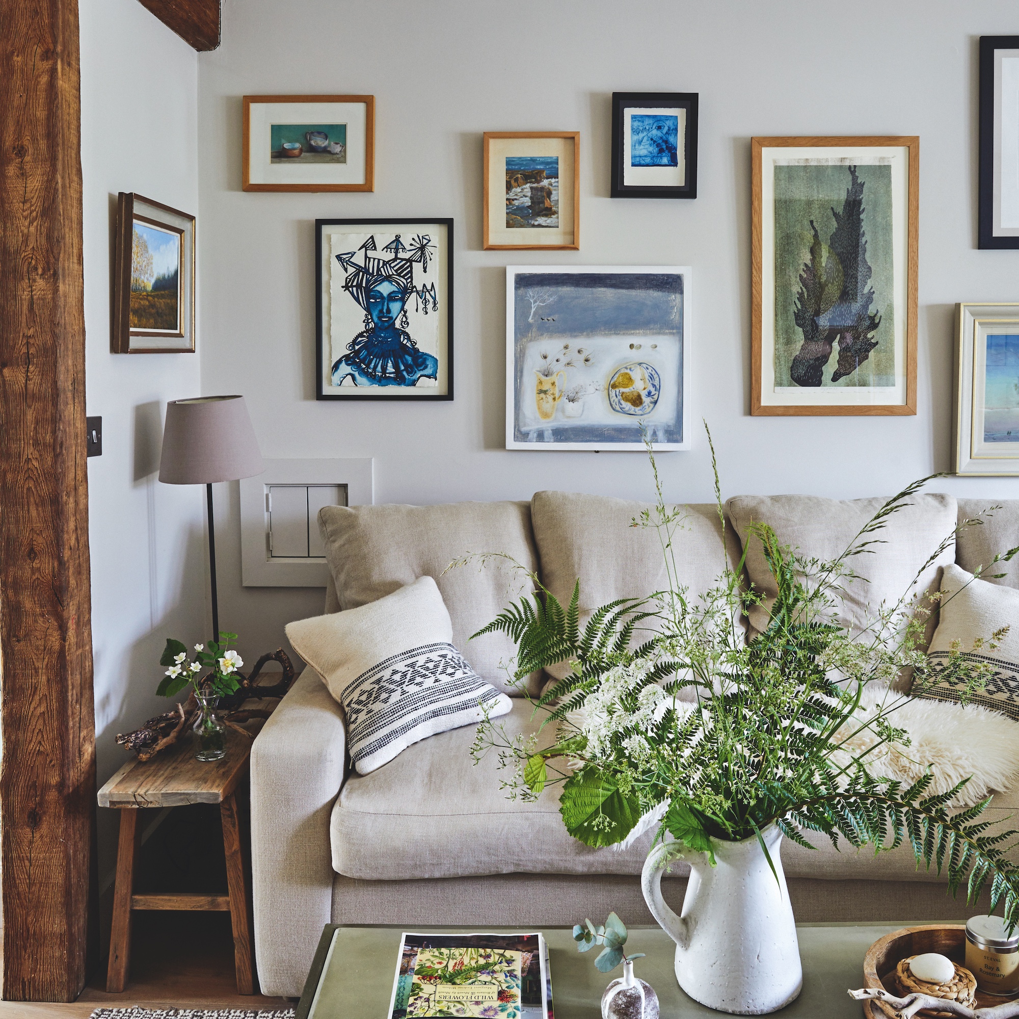 sitting room with cream sofa and gallery wall above of original paintings, with coffee table with jug of foliage