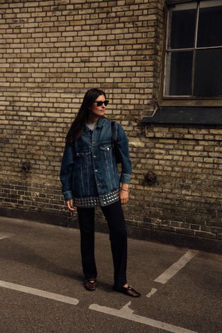 A guest attends copenhagen spring summer 2024 fashion week wearing a dark studded jean jacket, black pants, mesh flats and sunglasses