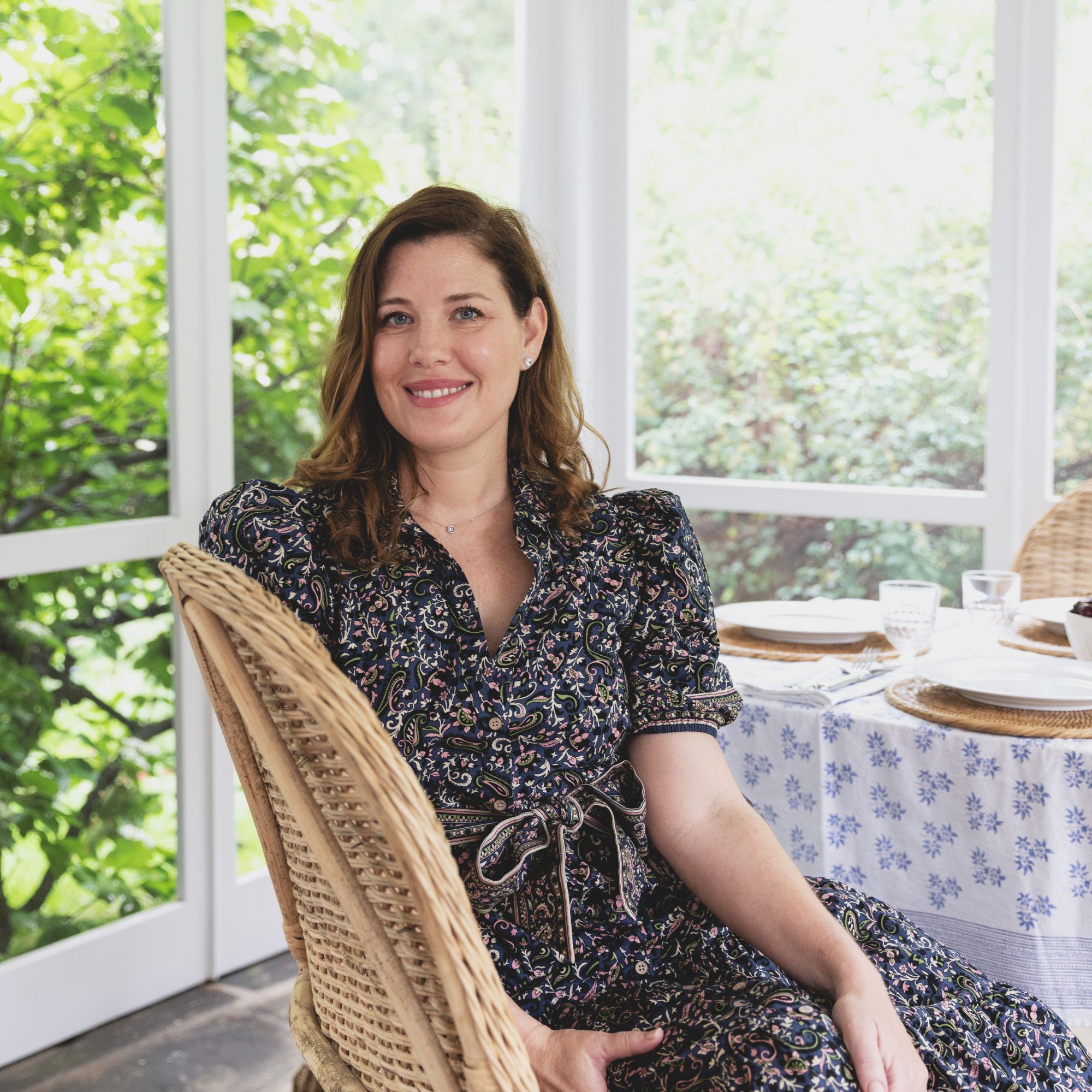 A picture of Jennifer Muirhead, a woman wearing a black dress sitting on a rattan chair in a conservatory