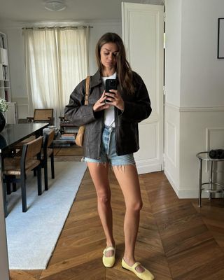 French female fashion influencer Anne Laure Mais poses for a mirror selfie wearing a collared leather jacket, tan shoulder bag, white T-shirt, cutoff denim shorts, and yellow Mary Janes flats