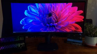 A curved Alienware AW3426DW widescreen monitor displays a vibrant flower image on a wooden desk, next to an open laptop with a lit keyboard, books, and a potted plant.