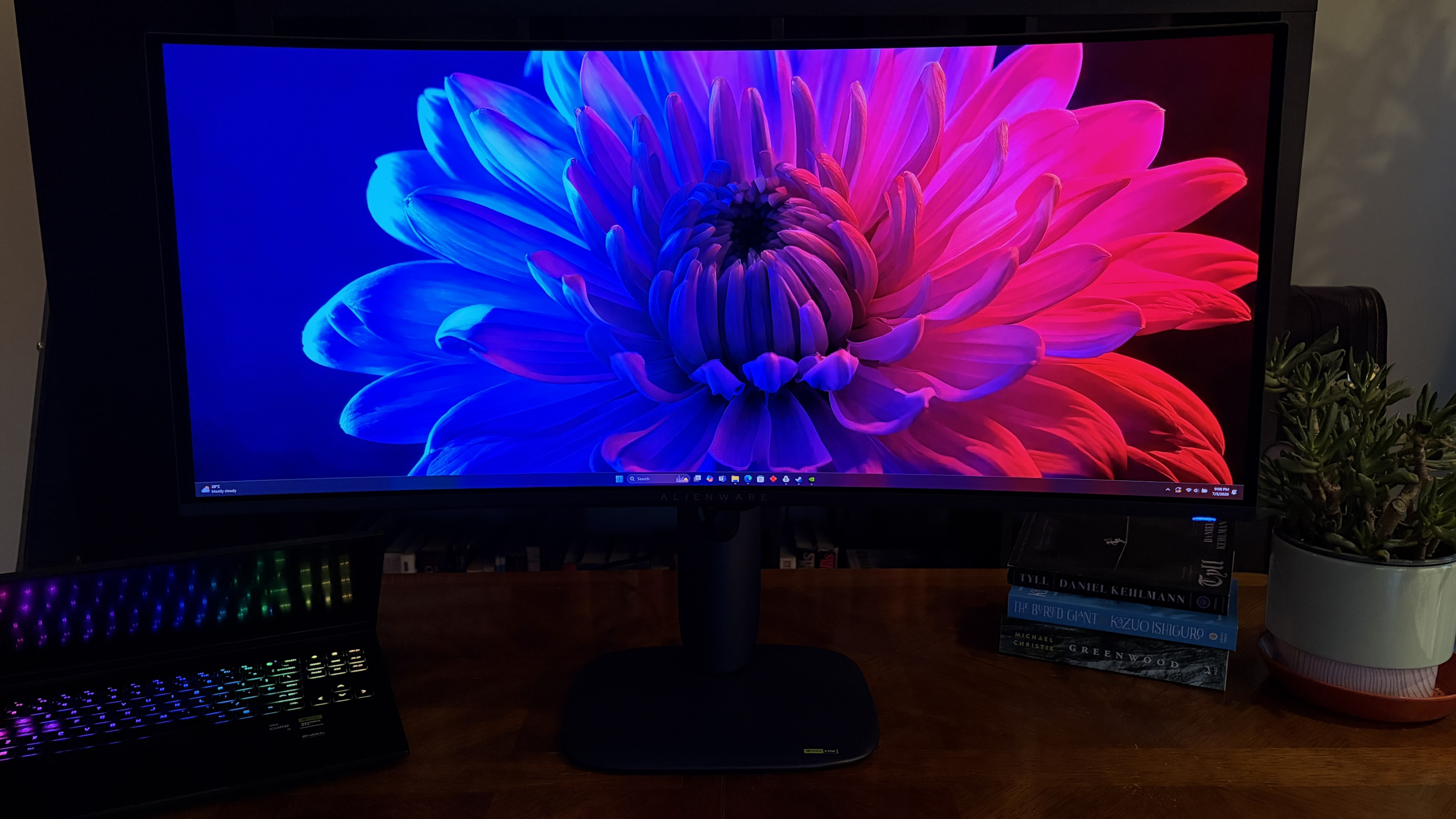 A curved Alienware AW3426DW widescreen monitor displays a vibrant flower image on a wooden desk, next to an open laptop with a lit keyboard, books, and a potted plant.