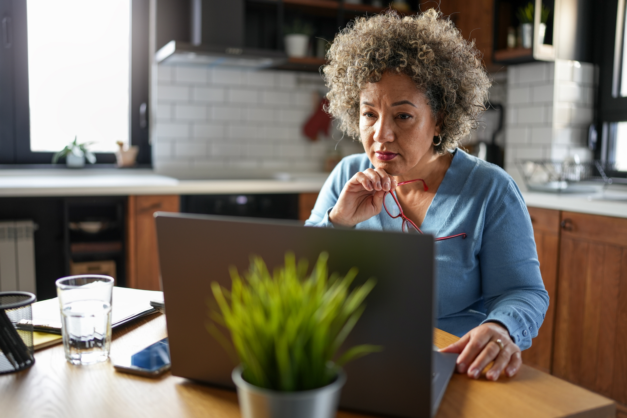A concerned mature woman with curly gray hair reviews paperwork at her kitchen table. She holds a document while looking at it intently, surrounded by a laptop, calculator, and papers