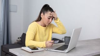 A woman with long brown hair in a ponytail looks down at her computer in a distressed manner. She is holding her forehead with one hand and a credit card with the other